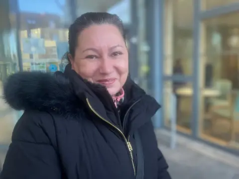 A smiling woman in a black coat with a fur hood stands outside a the glass doors of a building.