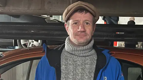 A man is standing next to an orange car. He is wearing a grey knitted jumper, a blue waterproof coat and a beige flat cap and is smiling at the camera. 