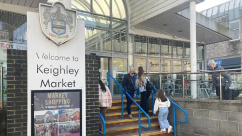 BBC A sign with 'Welcome to Keighley Market' and people climbing up some stairs under a covered entrance 