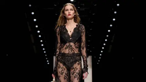 Getty Images A model walks the runway at the Dolce&Gabbana fashion show during the Milan Fashion Week - Womenswear Fall/Winter 2026/2027 on February 28, 2026 in Milan, Italy. She wears a black lace outfit with long light brown hair and bright red lipstick.