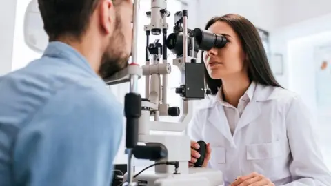 An eye test being carried out. We see the back of a man with dark hair and a short dark beard wearing a blue shirt. His chin rests on part of the eye test equipment. An optician with long dark hair and a white coat, looks through the eye piece of the equipment.