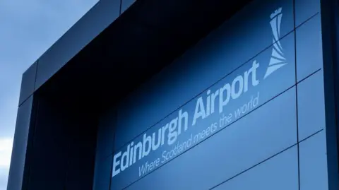 A general view of Edinburgh Airport. The name of the building is in white on a grey background. It is an overcast day.
