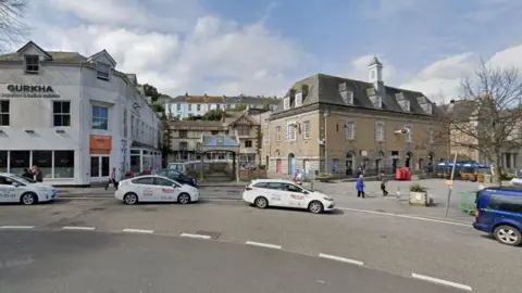 A small town square with white taxis parked outside. The square is made up of a white three storey building on the left, a beige coloured three storey building in the centre and a second three storey beige building on the right. 