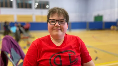 Jade sits in the middle of a gymnasium. She is wearing glasses and a red T-shirt and she is smiling at the camera. There are some chairs behind her.