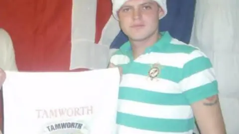 Scott Farrington A young Scott Farrington holding a Tamworth cloth. He is wearing a white and blue polo shirt and has a hat on. Half of his Tamworth FC tattoo can be seen. 