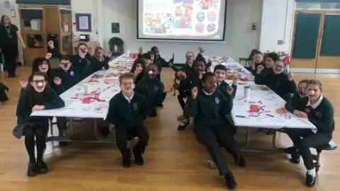 ALEXANDER STEVANOVIC/BBC About 20 schoolchildren are sitting in a school hall around two separate tables, wearing their own red noses. The tables have doodles on them and paint pens. The floor is a light coloured wooden floor