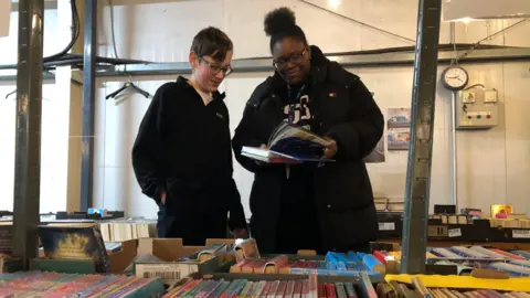 Pupil Evan with Miss Rowe-McIntosh who is looking through a book while they are both stood behind a table with books on it