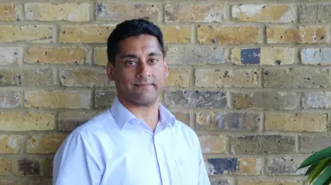 Raman Chaggar Raman Chaggar wearning a white shirt stands against a brick wall