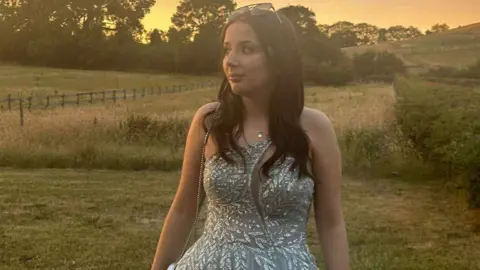 Family handout A girl in a dress stands in a field at sunset, with trees in the background.
