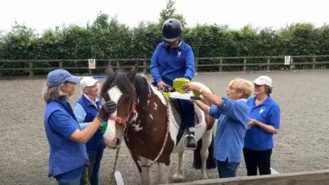 A group of volunteers stand around a client on a brown and white pony, one holds a device that the rider is interacting with