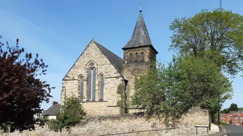 A stone-built church. It has a triangular-shaped roof with a spire on the right-hand side of the building. There are trees on either side of the building.