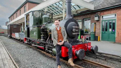 Nathan Spence Tim Dunn sits on the front buffer beam of a small green steam locomotive looking at the camera. Tywyn Wharf station is in the background