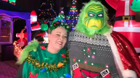 A woman in a christmas tree costume, and a man in a Grinch outfit smile for a photograph. Behind them are lots of inflated christmas decorations on their driveway