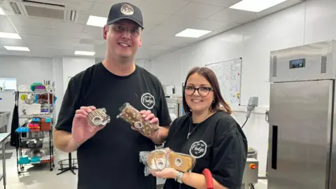 BBC Pete and Claire Brennan are pictured in their factory kitchen. They are both wearing black t-shirts featuring their company logo. Pete is also wearing a black baseball cap. Claire has dark hair and is wearing glasses. The couple are both holding a selection of fudge and chocolate products.