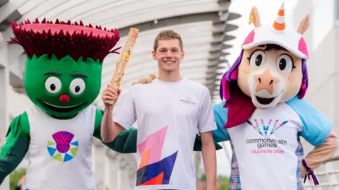 Getty Images Duncan Scott (centre) holding aloft a wooden Commonwealth Games baton. He is flanked by two mascots - Clyde, a green thistle, and a unicorn called Finnie, who has a traffic cone instead of a conventional horn. 