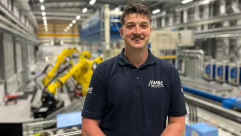 A man with short brown hair and a moustache is wearing a navy blue polo top with the words AMRC and smiling. He is standing in a huge factory with yellow machines in the background