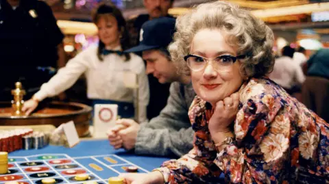 Caroline Aherne as Mrs Merton leaning over a blackjack table at casino in Las Vegas. She is leaning over the table, chips in hand, wearing a floral blouse, black glasses and silver wig. 