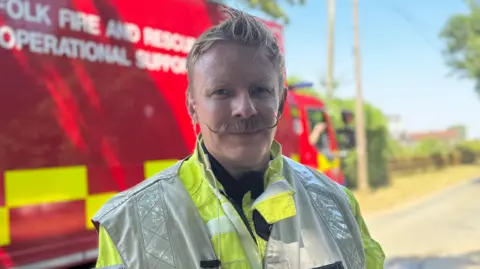 George King/BBC A firefighter stands in front of a fire engine. He has blond hair, a moustache and is wearing a bright green hi-vis jacket. 