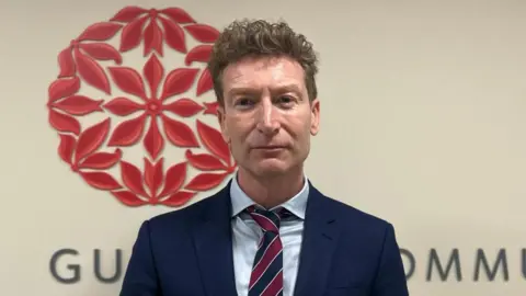Jim Roberts - A man with short brown hair, wearing a blue suit and red and blue tie, looks at the camera. he is standing in front of a wall with the flower-based logo of the Guernsey Community Foundation