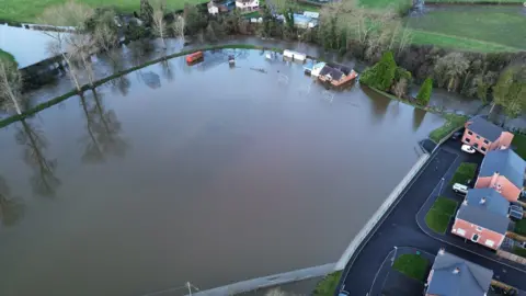 William Fisher An aerial shot of a cricket field that is totally covered in flood water. It is surrounded by roads on each side - on the road on the right are new red brick detached house. On the other sides are trees - a mixture of evergreen and deciduous. 