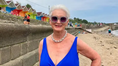 An older woman wearing diamanté sunglasses, a thick silver chain necklace and a bright blue swimming costume smiles at the camera. Behind her, a row of bright swimming hut chalets can be seen.