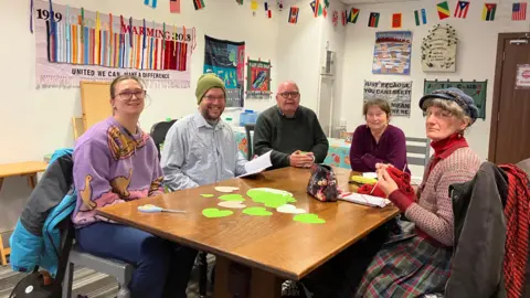 Jon Wright/BBC Five people sit round a large wooden table with cut out green paper hearts. There are banners on the walls.