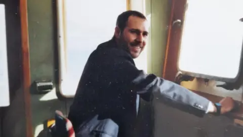 Family Handout A younger Francesco Galia is pictured on a ship. He is sitting down and smiling at the camera. He is wearing a dark suit jacket and has dark brown hair and beard.