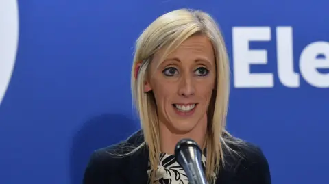 PA Media Carla Lockhart. She has blonde shoulder-length blonde hair, wearing a black blazer and black and white top.