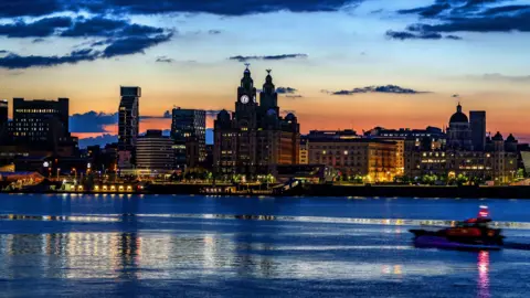 PA Media Liverpool’s waterfront at dusk, with the iconic Royal Liver Building and surrounding architecture illuminated against the sunset. The River Mersey reflects the city lights, while a blurred boat passes in the foreground.