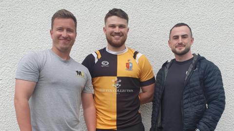 Head coach Matty Kaye, senior player and assistant coach Huw Evans and club physio Nathan Woods standing, smiling, in front of a pebble-dashed wall.