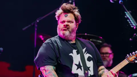 Depot Live Jaret Reddick is pictured whilst performing on stage at Cardiff Castle. He is wearing a black t-shirt with a white graphic print in the centre of it. He has brown hair, tattoos on his arms, and is holding a guitar. On the middle of his forehead is a guitar pick.