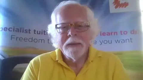 John Rogers, Chief Executive of the Association of Disability Driving Instructors, He is wearing a yellow polo shirt and has glasses, grey hair and a grey goatee.