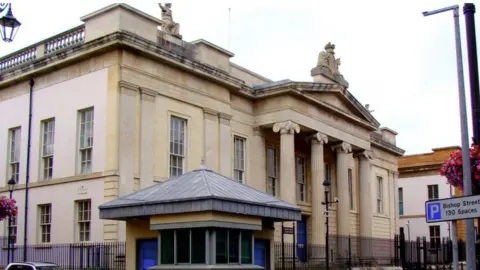 Shows a large court building with four pillars at the front, a security hut to the left and a silver-coloured car to the extreme left. 