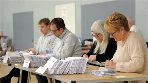 BBC Two dark haired men in grey jumpers and two women, one with white hair, a black top and glasses and one with blonde short hair and a cream jumper counting ballot papers at a fold-down table, with three baskets of ballots in front of them