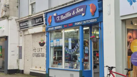 The T Baillie and Son butcher shop - it is a small shop with blue storefront. Bunting saying Retirement is hanging in the window.