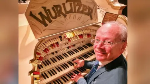 Phil Kelsall sits at the console of the Wurlitzer organ in Blackpool Tower Ballroom with his hands on the keyboards. He's wearing a dark grey suit, white shirt and blue tie.