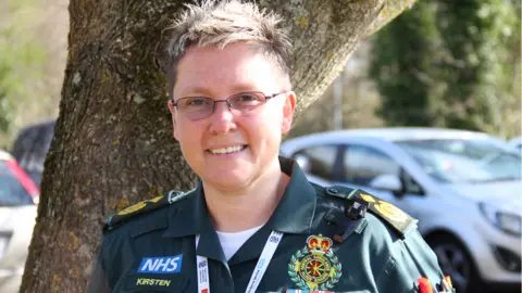 SCAS Kirsten Willis-Drewett is wearing her ambulance uniform and is stood in front of a couple of cars. She is wearing glasses and has short, blonde hair.