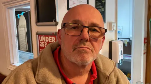 Bene has a bald head and black rimmed glasses. He wears a light brown fleece.