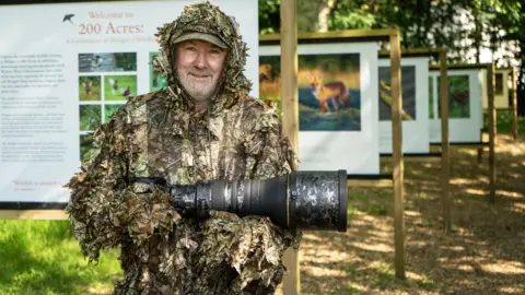 The Lost Gardens of Heligan Andy Wilson holding his camera which has a large telescopic lens attached. He is wearing a camouflage ghillie suit and a camouflage cap.