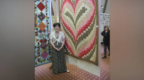 Rod Parsons Julie is stood in front of one of her quilts which is hanging behind her. It has a heart design and is pink, brown and cream. She is wearing a floral dress and and a white shawl.