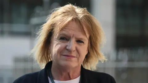 Jamie Niblock/BBC Deborah Coles has shoulder-length blonde hair and is wearing a black jumper over a white T-shirt. She is grimacing in front of a building in London.
