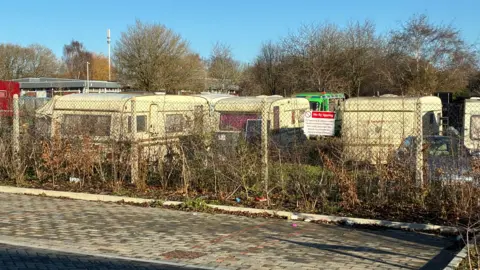 A row of caravans behind a wire fence, with some bushes in the foreground.
