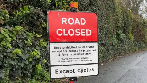 A road closed sign in Guernsey in front of a bush. It is split into three sections, The top section is red with "road closed" written on it. The section below is white and explains traffic cannot use the road in black letters. The lowest section says "except cycles" on a white background with black letters.
