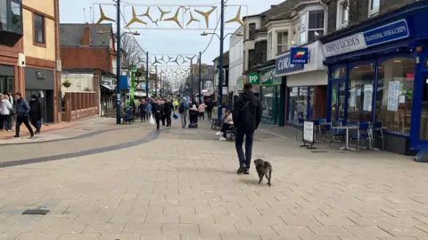 Owen Sennitt/BBC A man walks along Regent Road in Great Yarmouth, with his small dog following off-lead, as crowds of people walk down the street ahead of him