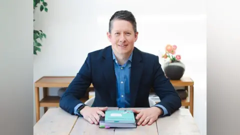 John Adams has brown hair slicked and styled to the side. He is sitting at a light wood table and is wearing a navy suit jacket and blur shirt. There is a blue book and a notebook in front of him.