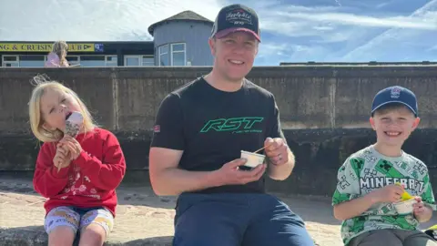 Daniel Ingham sitting on a wall by the beach below Peel Sailing Club with his young son and daughter either side of him. They are all smiling while eating ice cream.
