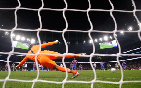 Reuters View through goal net, goalkeeper in orange jersey dives to left as ball from striker goes to the right from a striker in blue and purple