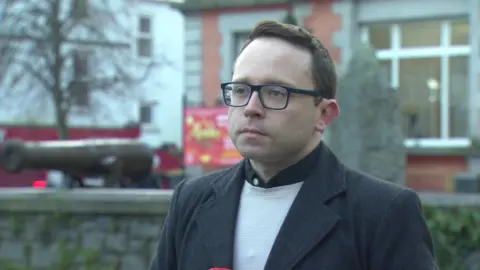 BBC News Dáire Hughes is looking off to the side of the camera. He has brown hair cut short. He is wearing dark rimmed glasses, a black wool coat and a cream jumper. His expression is solemn. Behind him is a stone wall with a cannon on top, as well as a red-brick building.