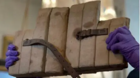 A person with purple rubber gloves on holds up a cork life jacket designed in the 1800s in Cornwall. It has a brown leather belt attached to it.