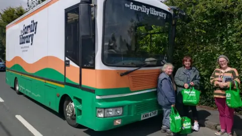 The Family Library A large green, orange and white bus is parked on a road. Three women with green bags, that read mobile library, stand in front of it.